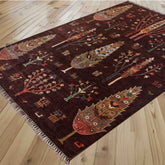 A dark brown area rug with a detailed, colourful tribal tree pattern sprinkled with stylised animals and birds, laid out on a light-colored wooden floor.