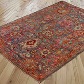 A rectangular area rug with a traditional floral pattern in sage green, rose pink, heather, and terracotta, shown laid out on a light-colored wooden floor.