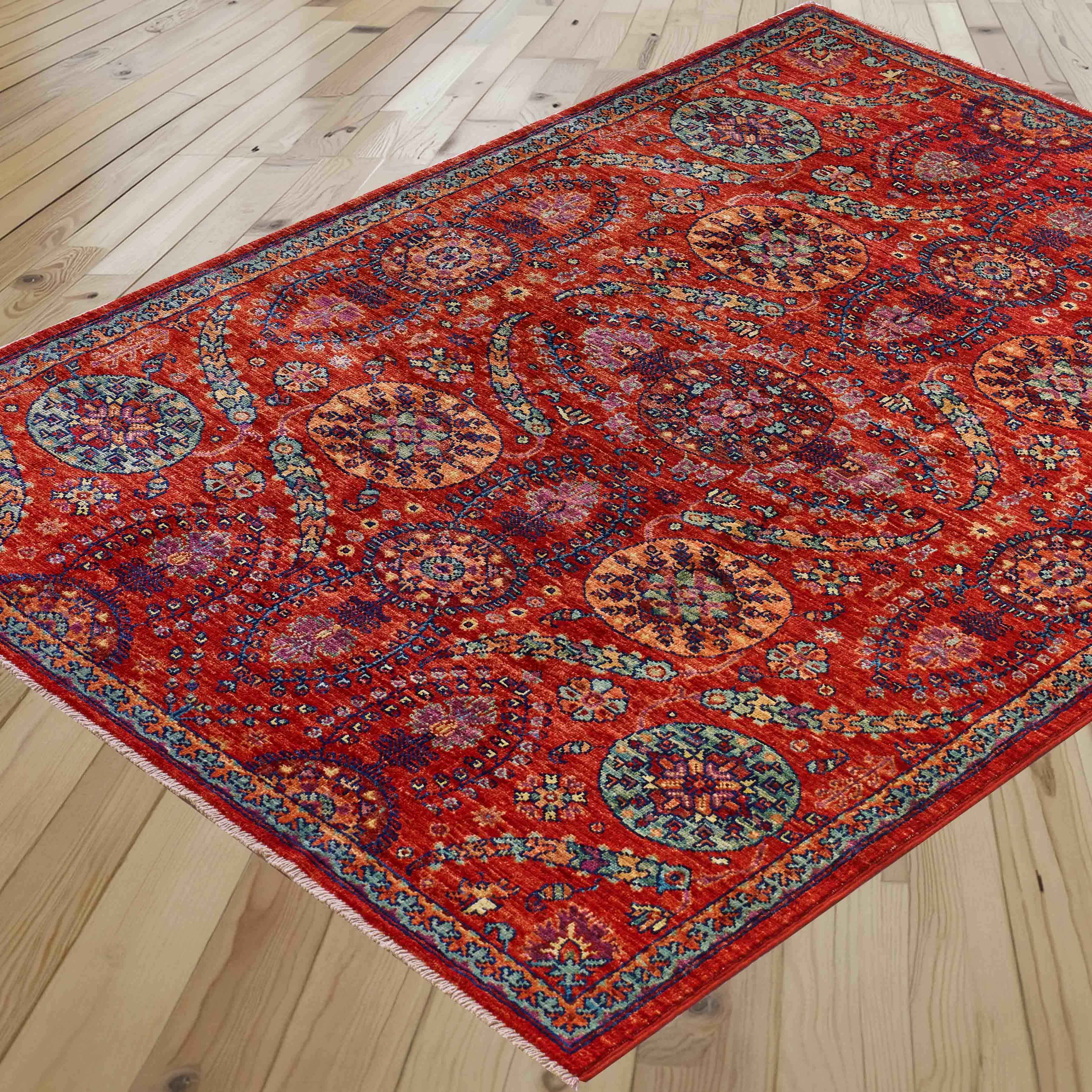 A traditional area rug with a vibrant red base and intricate multicolour floral patterns, shown laid out on a light wood plank floor.